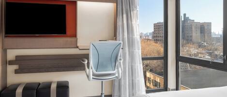 A hotel room with a large window, a flat-screen TV mounted on the wall, a modern office chair, and a bed.
