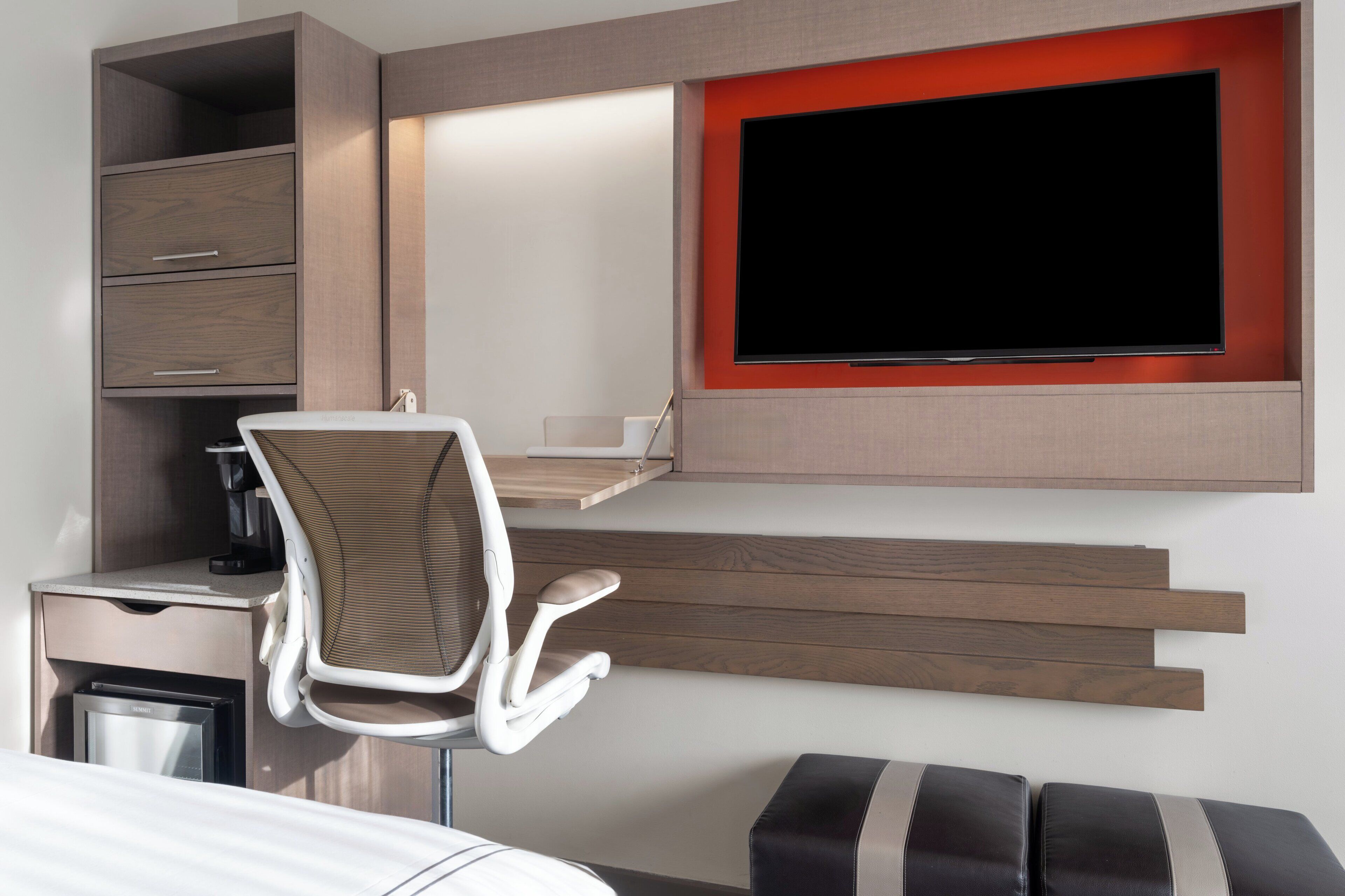 A modern hotel room with a desk, chair, flat-screen TV, built-in shelving, and a small kitchenette.