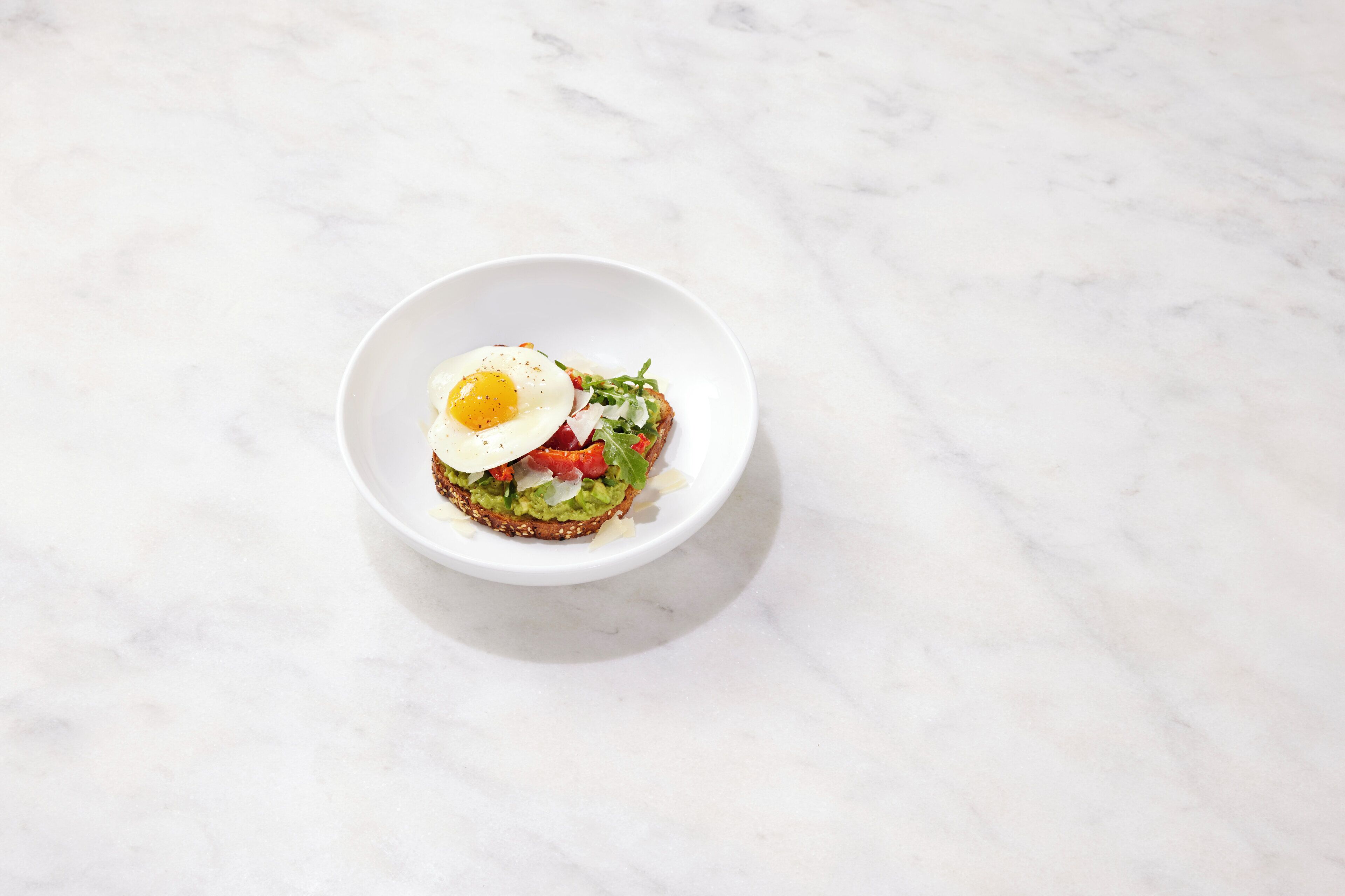 A white bowl containing a breakfast sandwich with an egg, avocado, tomato, and greens on a slice of bread, placed on a marble surface.