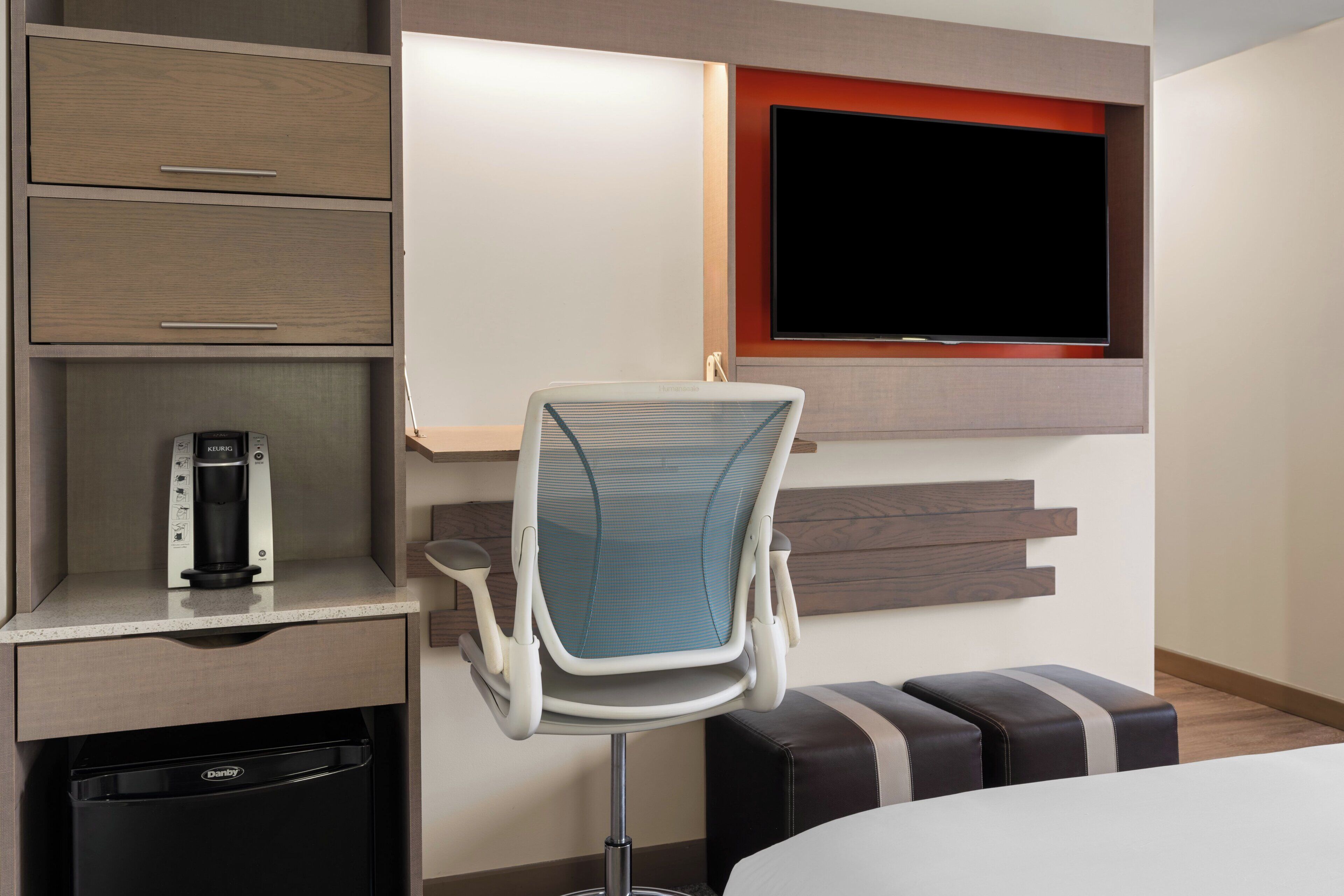 A hotel room with a bed, a desk with a coffee maker, a chair, a TV mounted on the wall, and a small trash bin.