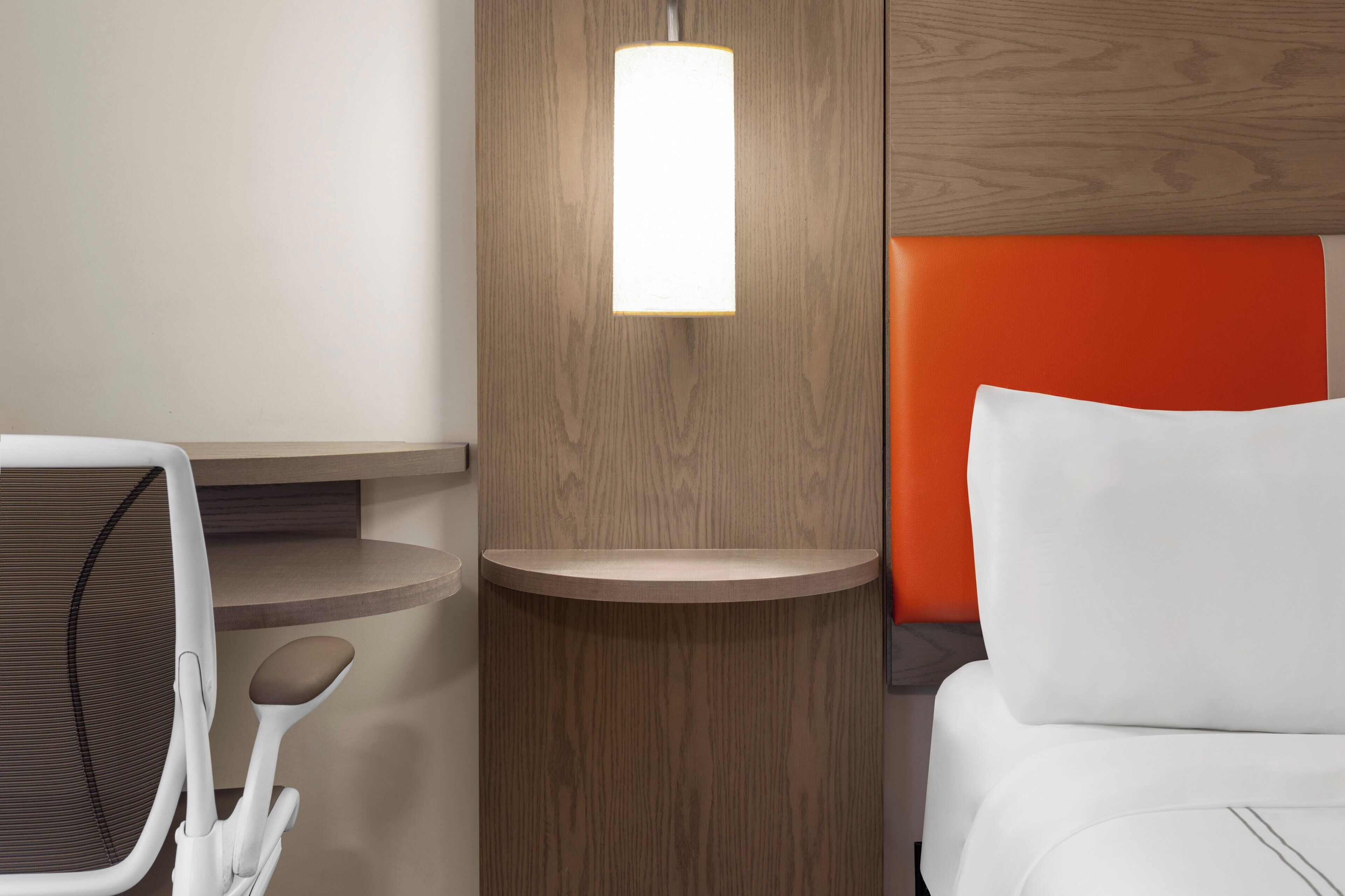 A hotel room with a bed, a desk, a chair, and a wall-mounted light fixture.