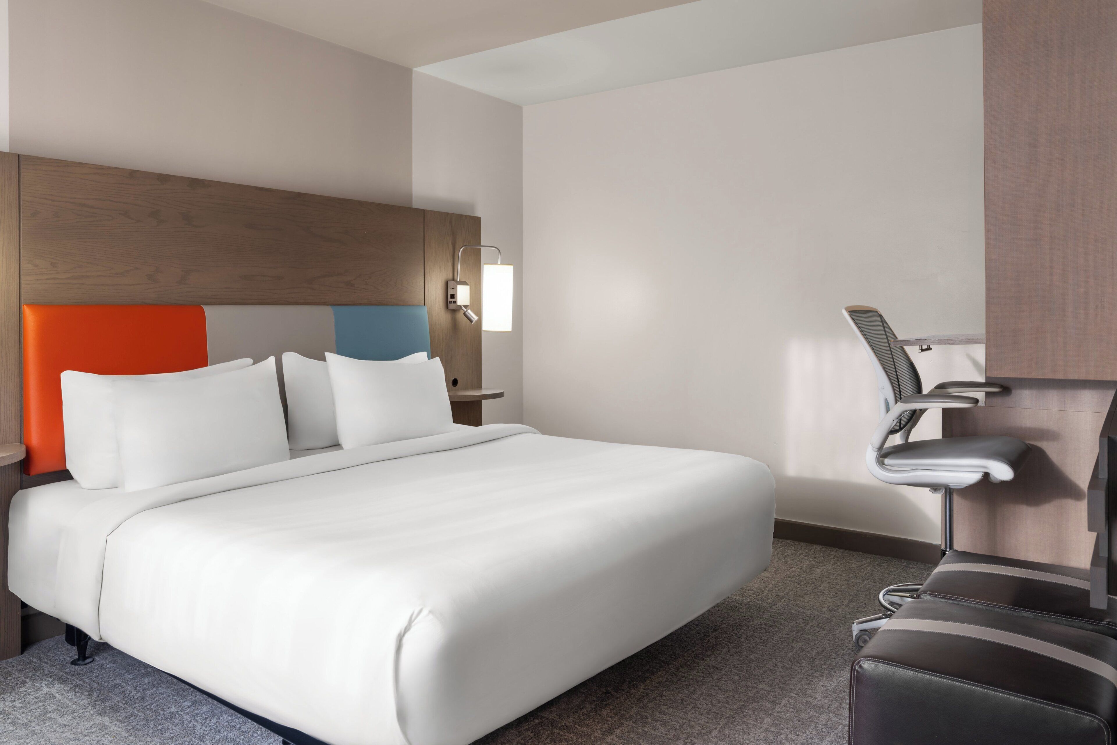 A hotel room with a large bed, a desk with a chair, and a wall-mounted light.