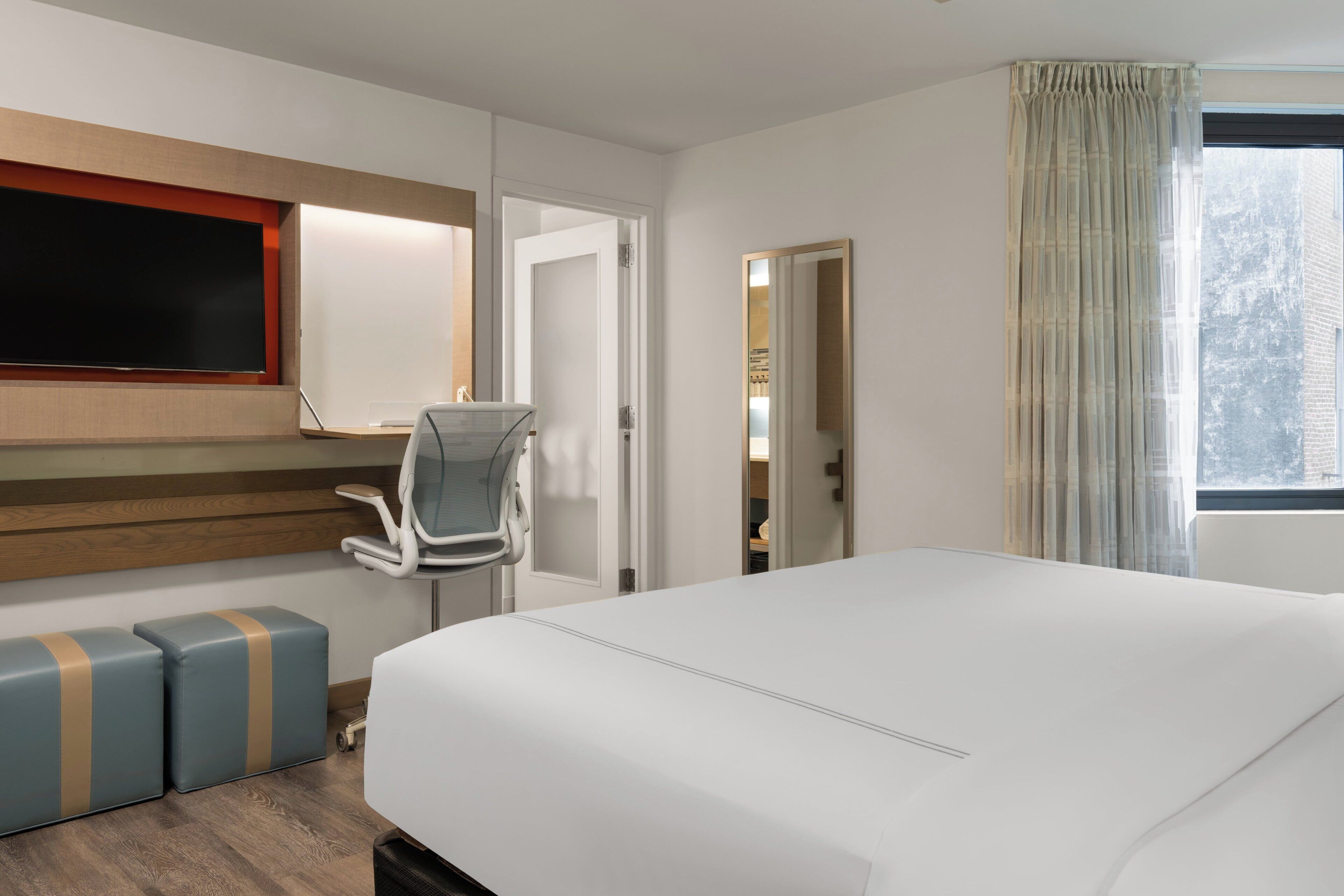 Habitación de hotel con una cama grande, un televisor montado en la pared, un escritorio con una silla y una ventana con cortinas.