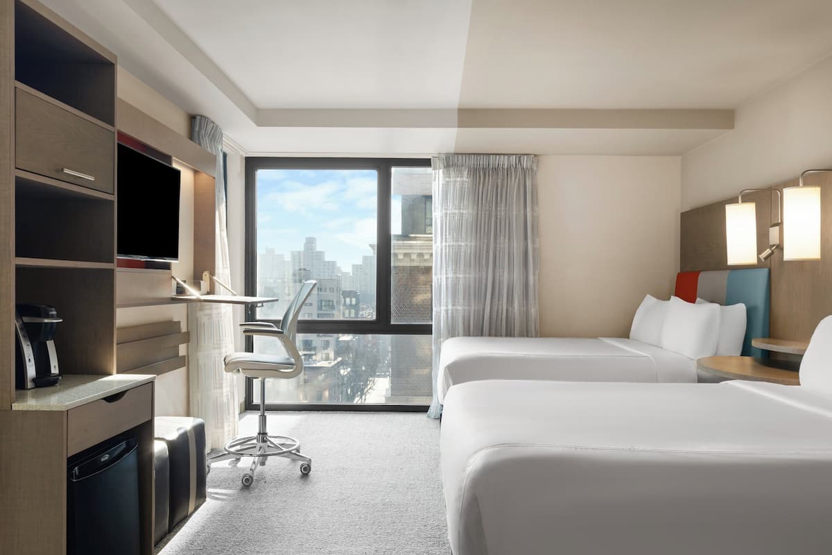 A hotel room with two beds, a desk, a chair, a coffee machine, and a large window with a city view.
