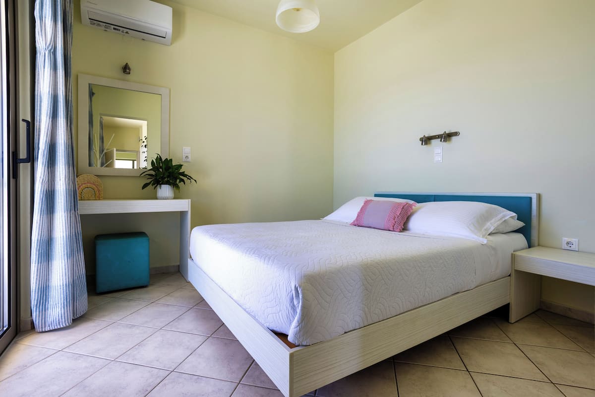 A bedroom with a bed, a bedside table, a nightstand, a mirror, a potted plant, and a window with curtains.