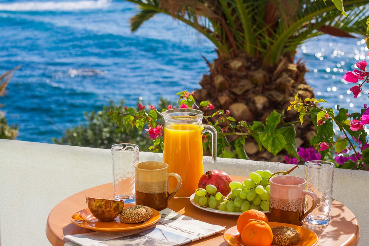 Un desayuno con un vaso de zumo de naranja, una taza de café, un plato de uvas, una naranja y una granada en una terraza con vistas al mar.
