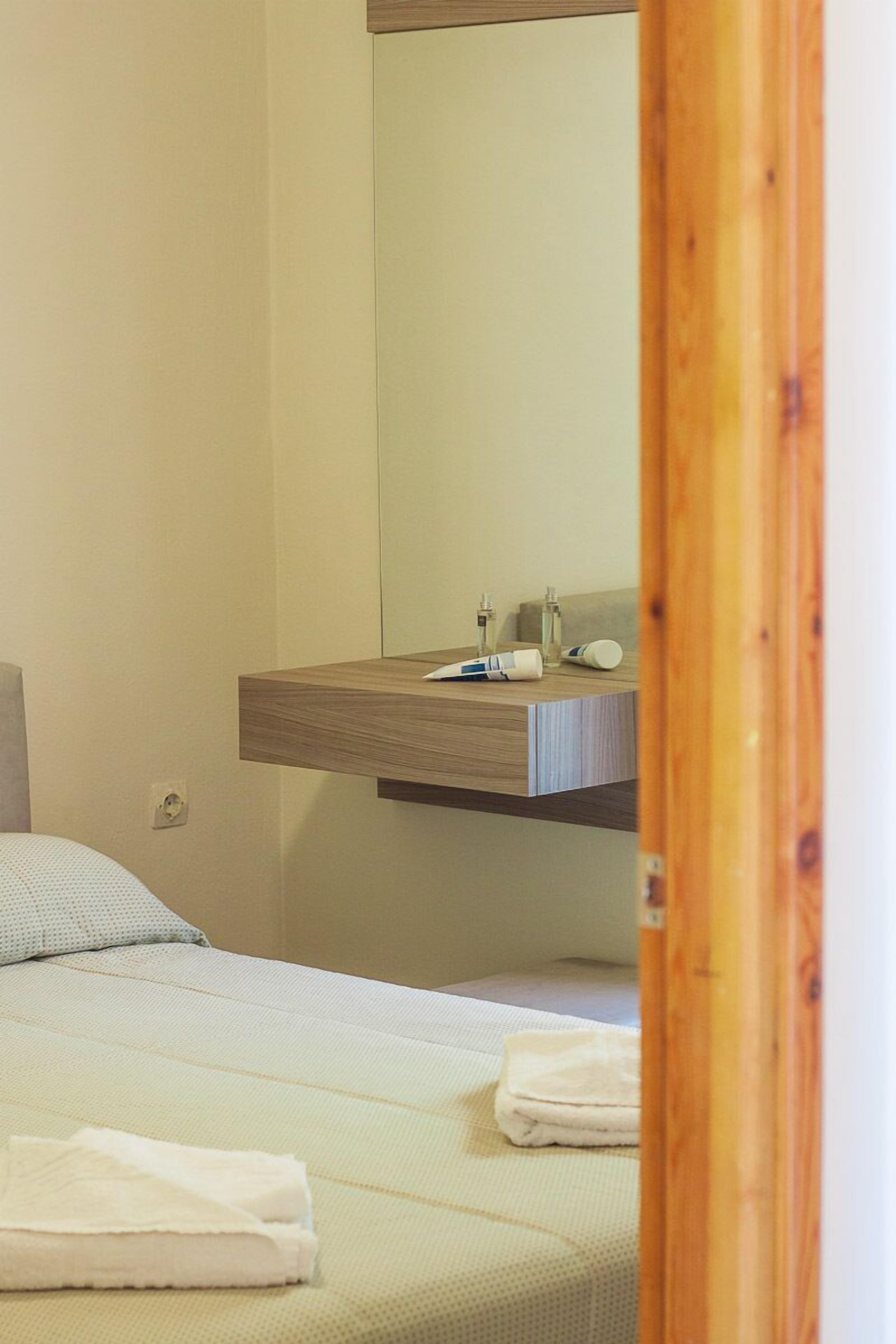 Una habitación de hotel con cama, mesita de noche de madera, espejo y un baño pequeño con lavabo y artículos de aseo.