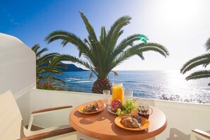A balcony with a table set for breakfast, overlooking a coastal view with palm trees and the sea. - Skinos Apartments and Studios (Agios Vasileios)