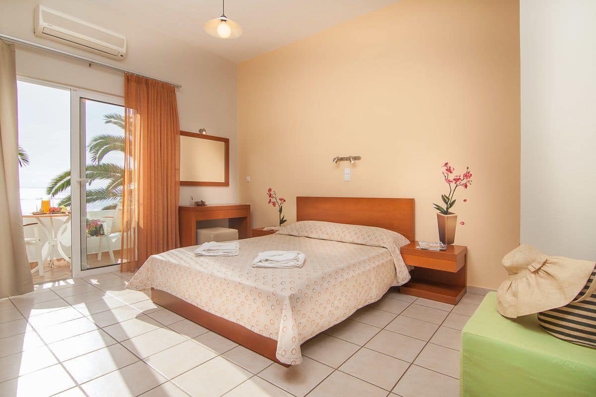 A hotel room with a bed, a nightstand, a green sofa, a potted plant, and a sliding glass door leading to an outdoor area with a table and chairs.