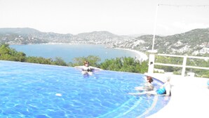 Une piscine à l’eau d’un bleu cristallin, entourée de verdure et offrant une vue sur un paysage côtier avec des collines et une plage.