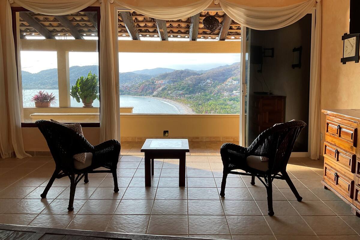 Une pièce avec vue sur une rivière et des collines, deux fauteuils en osier, une petite table et un buffet en bois.