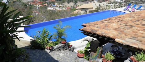 A swimming pool with a tiled roof, surrounded by potted plants and overlooking a residential area with houses and hills.