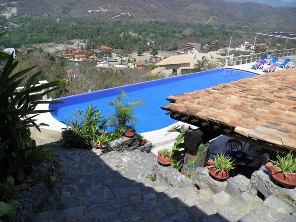 Una piscina con techo de azulejos, rodeada de plantas en macetas y con vistas a una zona residencial con casas y colinas.