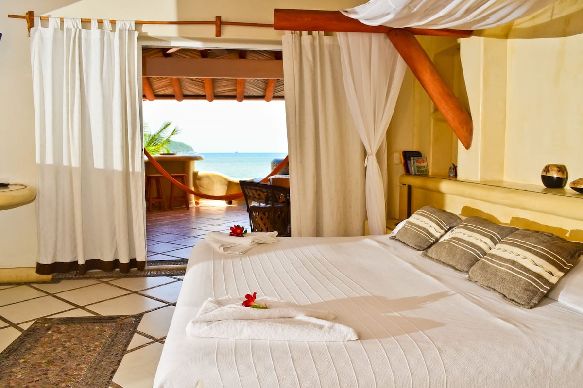 A neatly made bed with pillows and a towel, a small table with a chair, and a view of the sea through a doorway.