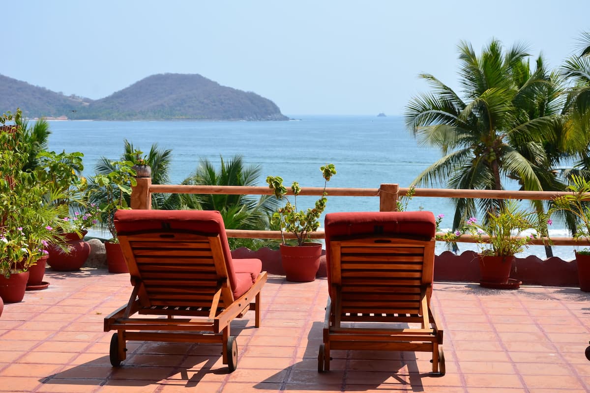 Two wooden lounge chairs with red cushions on a tiled patio overlooking a body of water and a distant mountain.