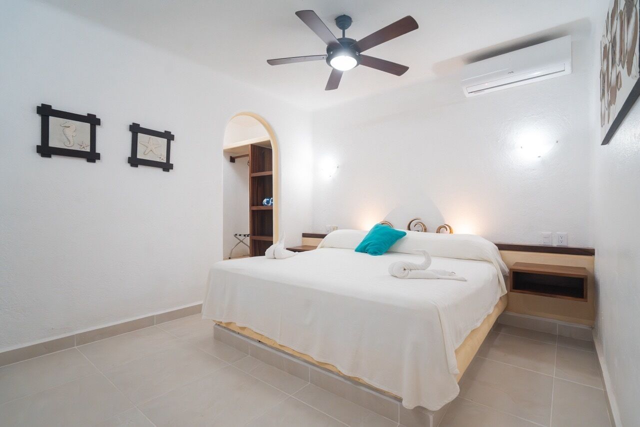 A bedroom with a bed, a ceiling fan, an air conditioning unit, and a small bedside table.