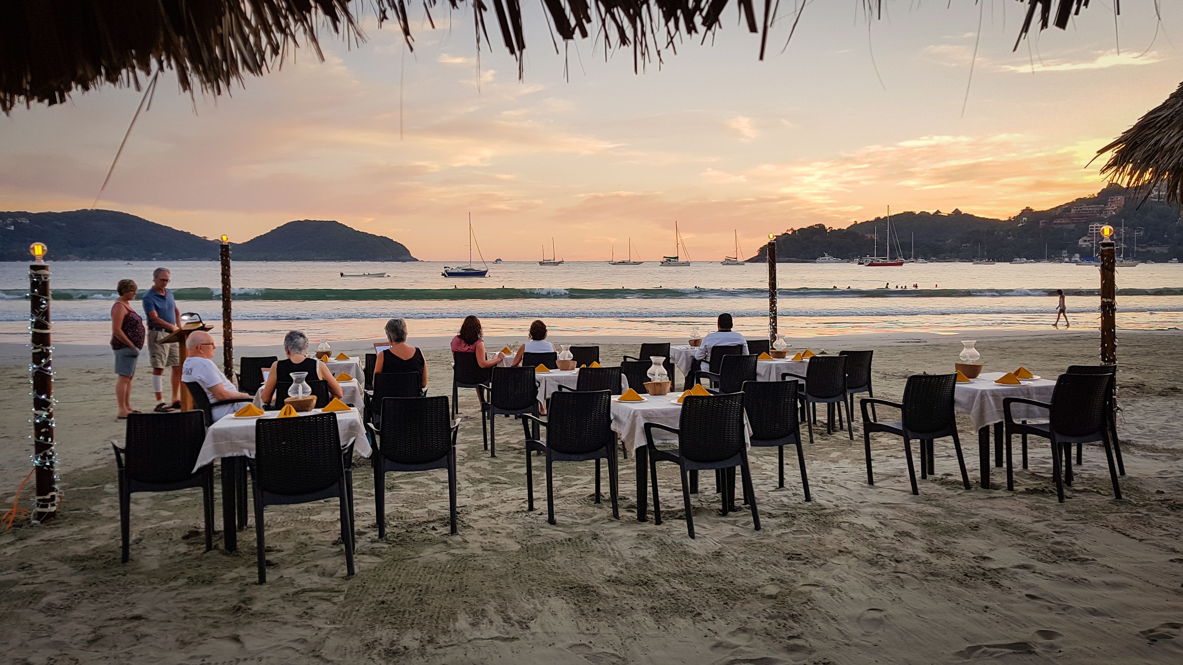 A beachside restaurant with tables and chairs arranged for dining, overlooking the ocean with boats and distant hills.