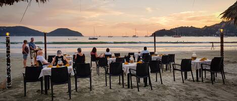 A beachside restaurant with tables and chairs arranged for dining, overlooking the ocean with boats and distant hills.