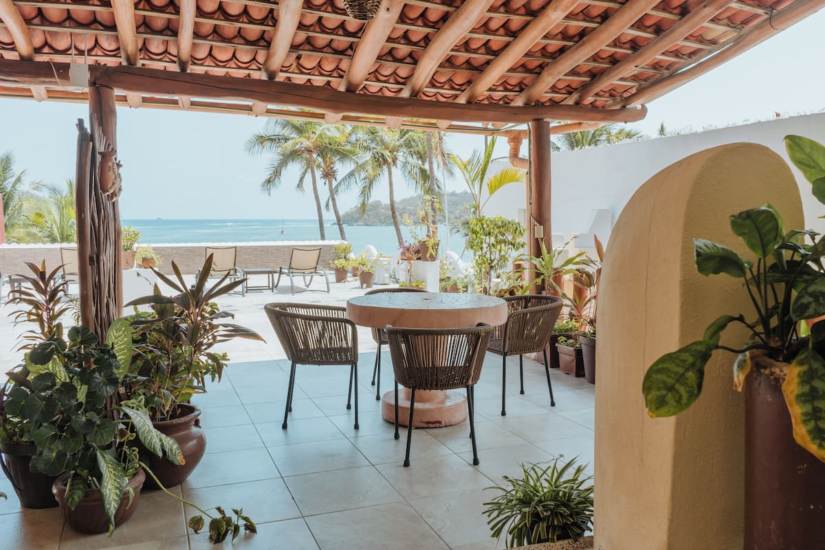 A covered patio with a round table and chairs, surrounded by potted plants, overlooking a beach with palm trees and the ocean.