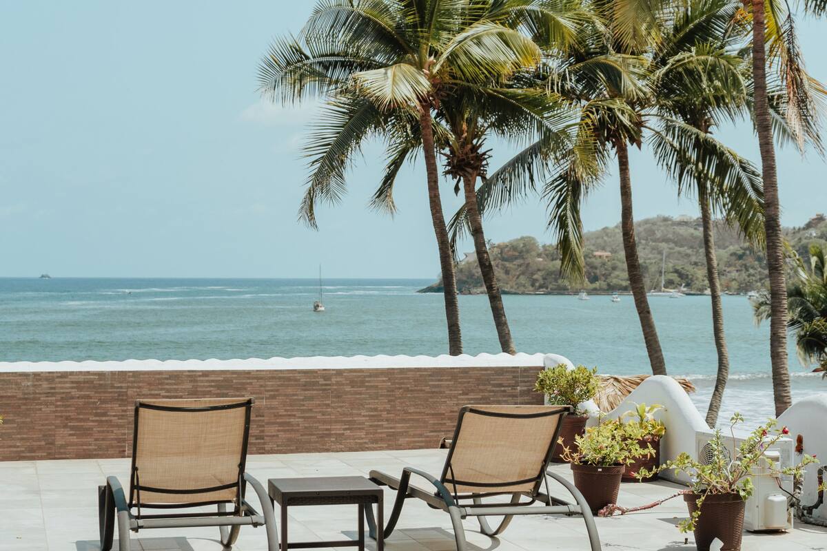 A beachside lounge with two lounge chairs and a small table, palm trees, and a view of the ocean.