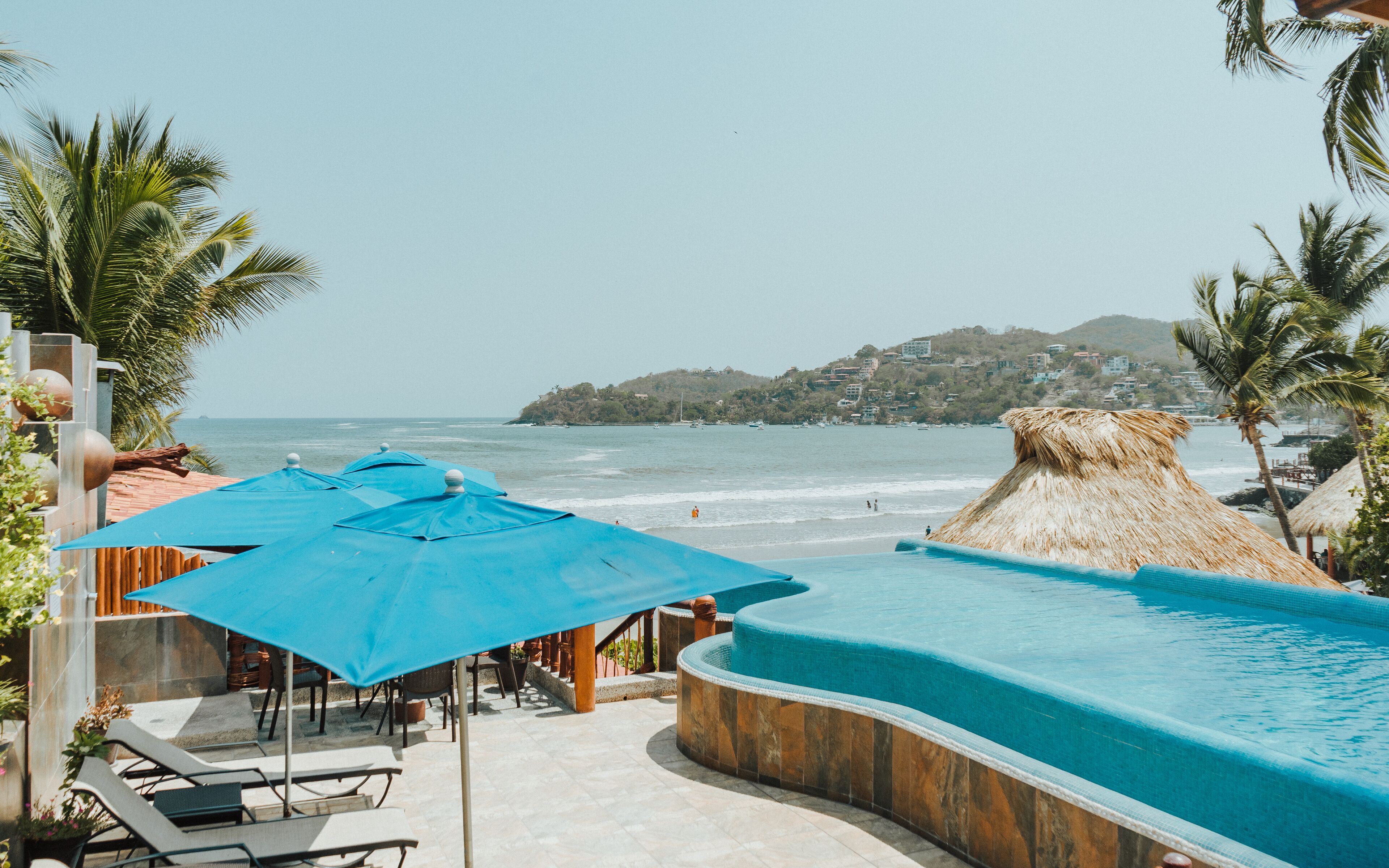 Un espace piscine agrémenté de parasols bleus, de chaises longues et offrant une vue sur la plage et l’océan.