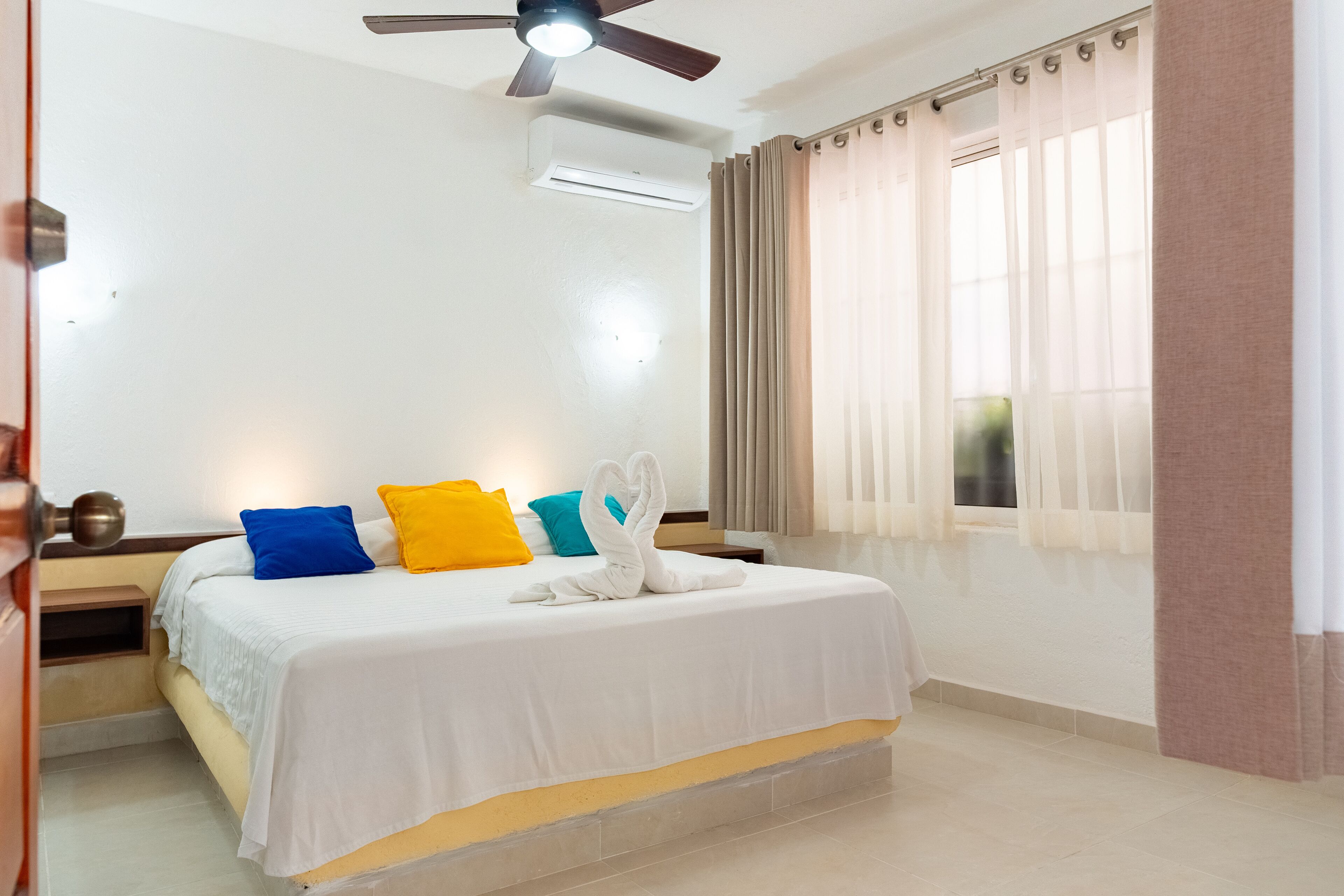 Una camera d'albergo con un letto, una ventola a soffitto, un condizionatore d'aria e una finestra con tende.