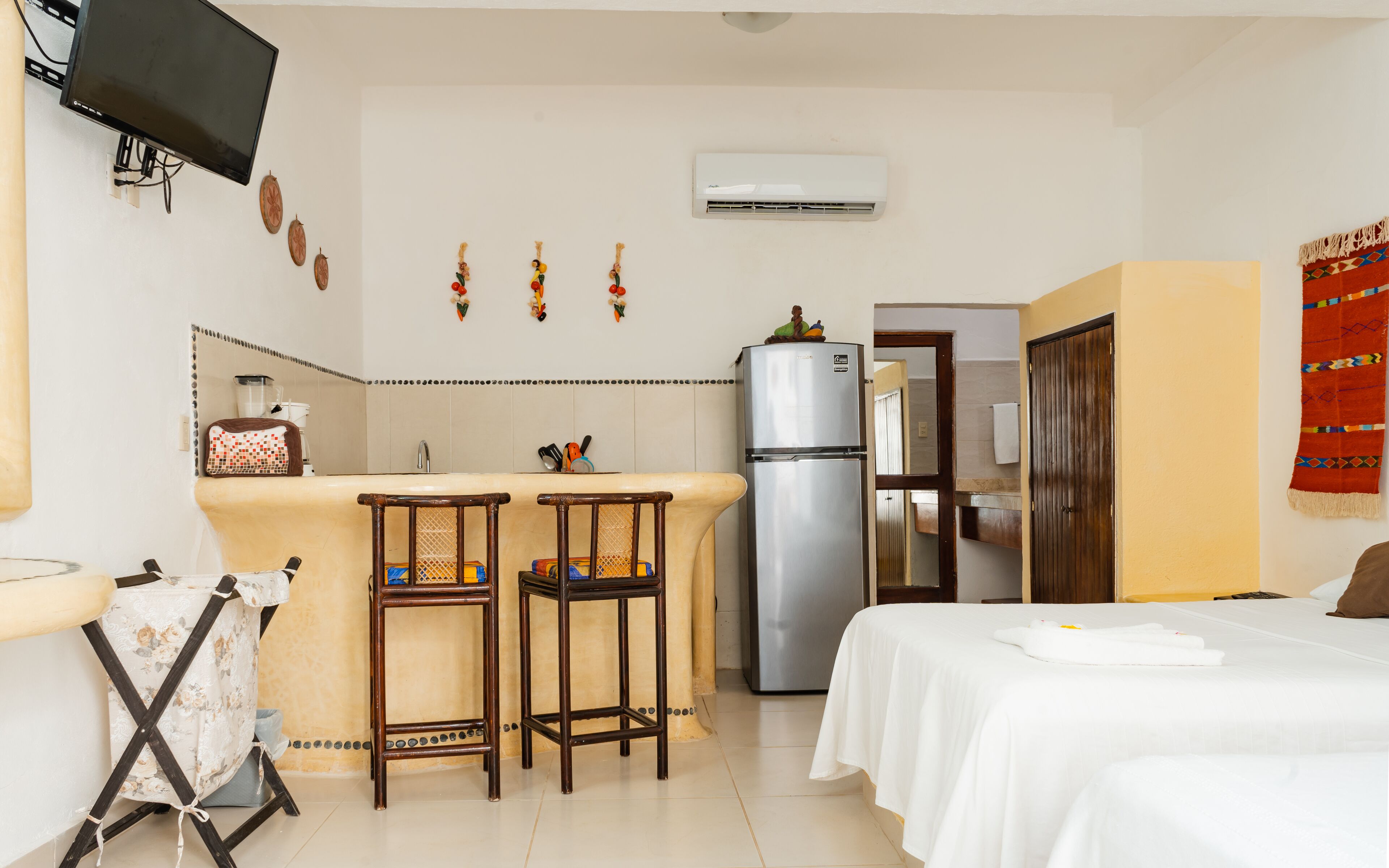 A compact hotel room with a kitchenette, a bed, and a small sitting area with two stools.