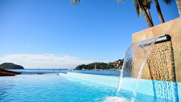 Une piscine à débordement avec une cascade, une eau d’un bleu cristallin et une vue sur la mer, où l’on peut apercevoir des bateaux.