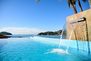 A infinity pool with a waterfall feature, clear blue water, and a view of the sea with boats. - Arena Suites (Zihuatanejo)