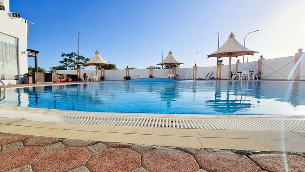 A swimming pool with lounge chairs and umbrellas, surrounded by a white building with a tiled deck.