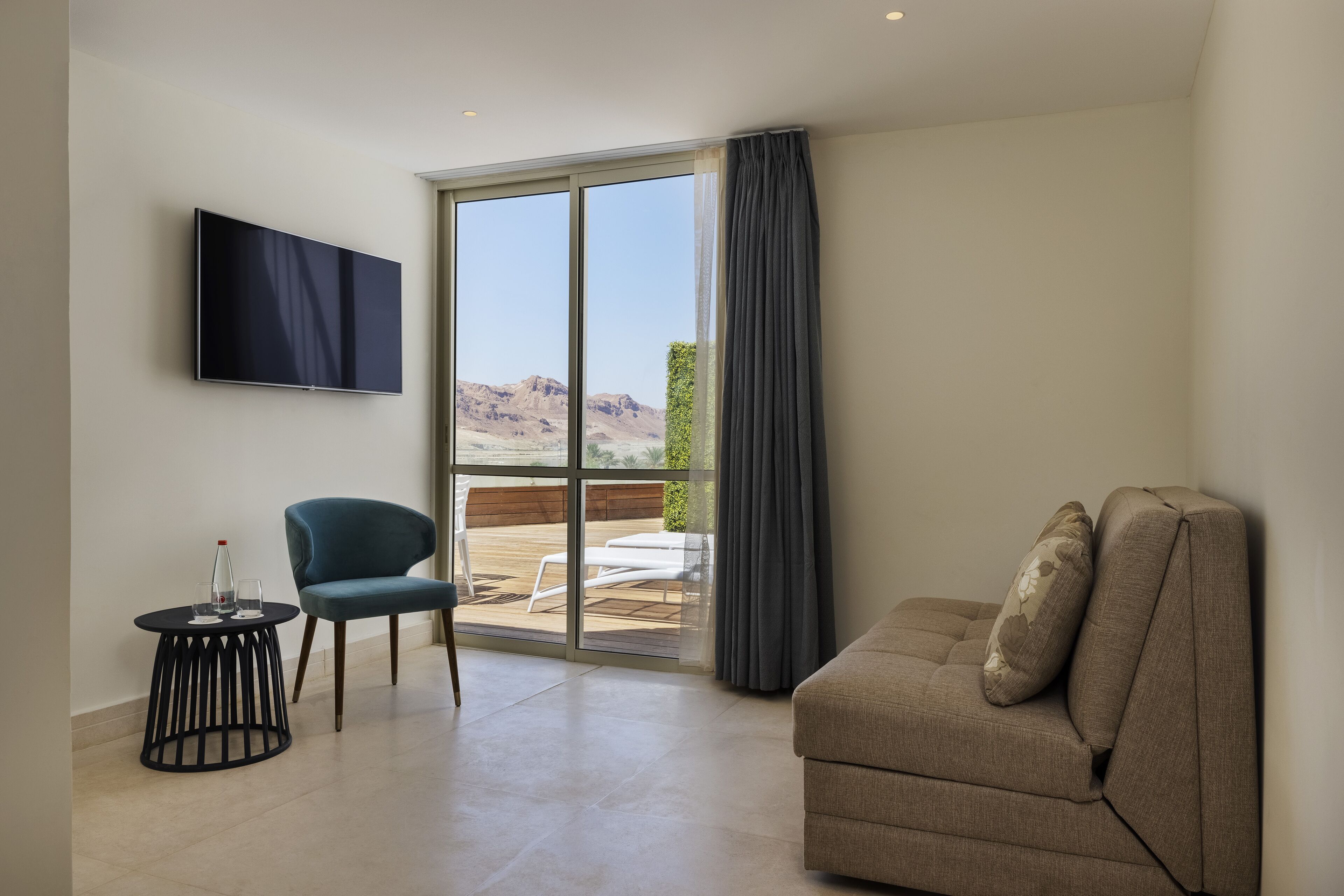A modern living room with a sofa, a chair, a TV mounted on the wall, and a sliding glass door leading to a balcony with a view of the mountains.