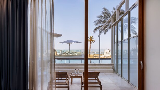 A room with a glass door leading to a balcony with a view of the sea and palm trees. - Herbert Samuel Hod Dead Sea Hotel (Tamar)