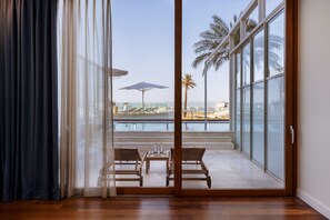 Una habitación con puerta de vidrio que da a un balcón con vista al mar y palmeras.