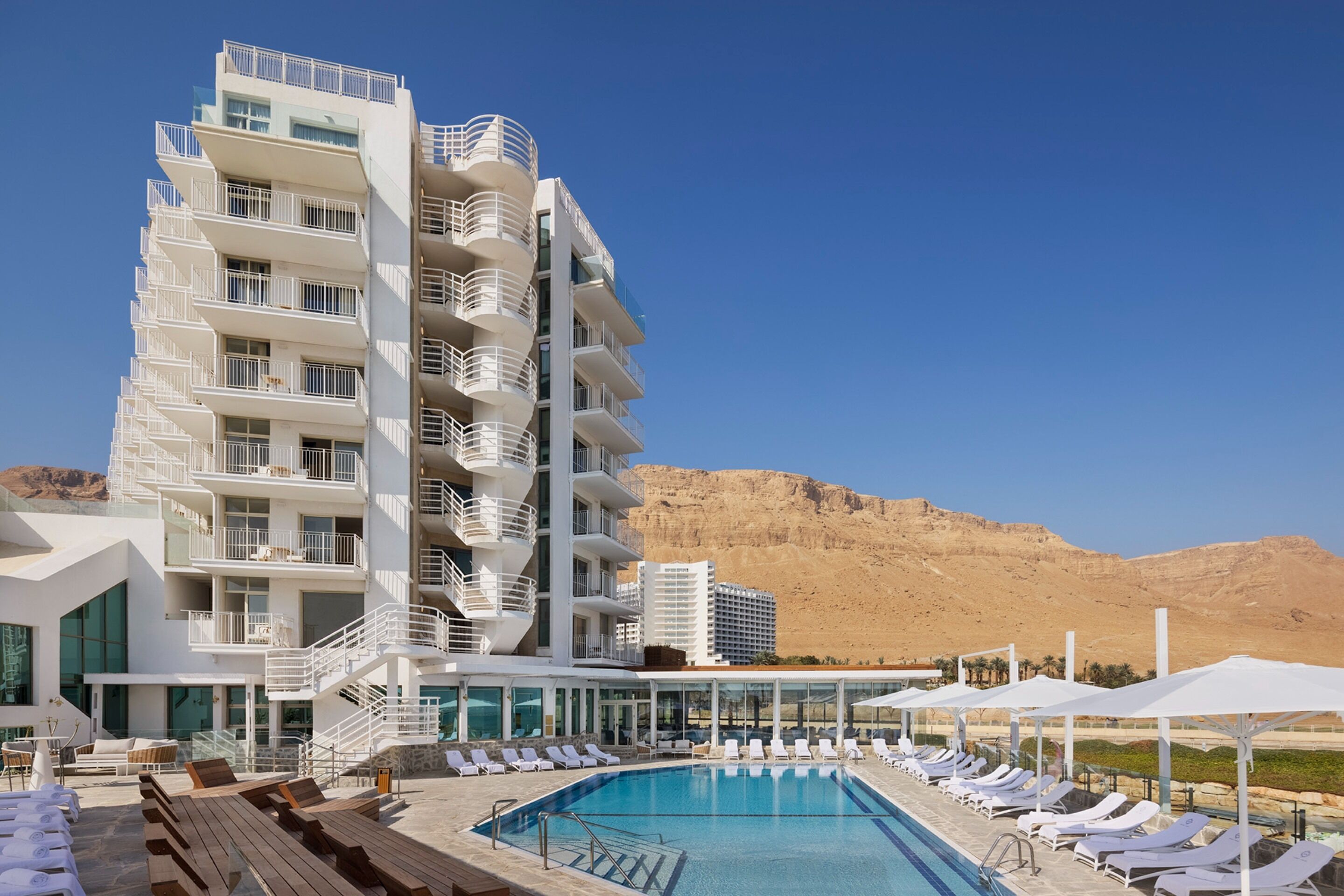 A modern hotel with a pool, sun loungers, and a view of a mountain.