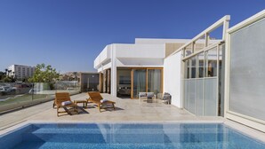 A modern pool area with two wooden lounge chairs and a small table, a glass-enclosed patio, and a clear view of the surrounding buildings. - Herbert Samuel Hod Dead Sea Hotel (Tamar)