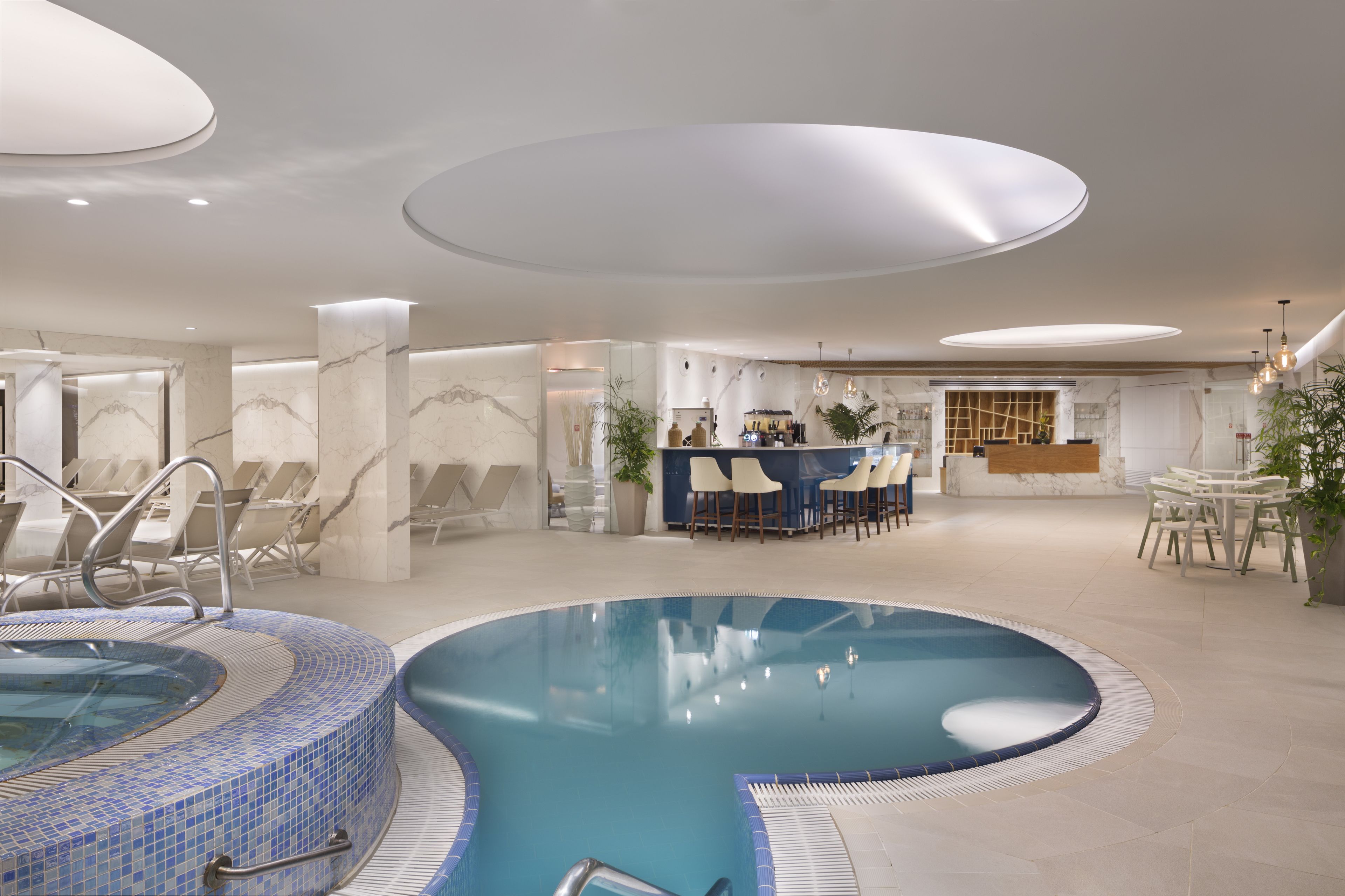 A modern indoor pool area with a hot tub, lounge chairs, and a reception desk.