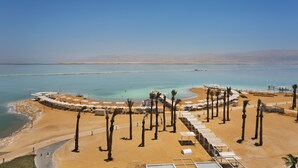 An aerial view of a beach resort with a circular layout of beach chairs and umbrellas, palm trees, and a clear view of the sea. - Herbert Samuel Hod Dead Sea Hotel (Tamar)