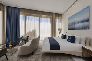 Una habitación de hotel moderna con una cama curvada, un sillón verde, una mesa de centro de vidrio y un ventanal con cortinas translúcidas.