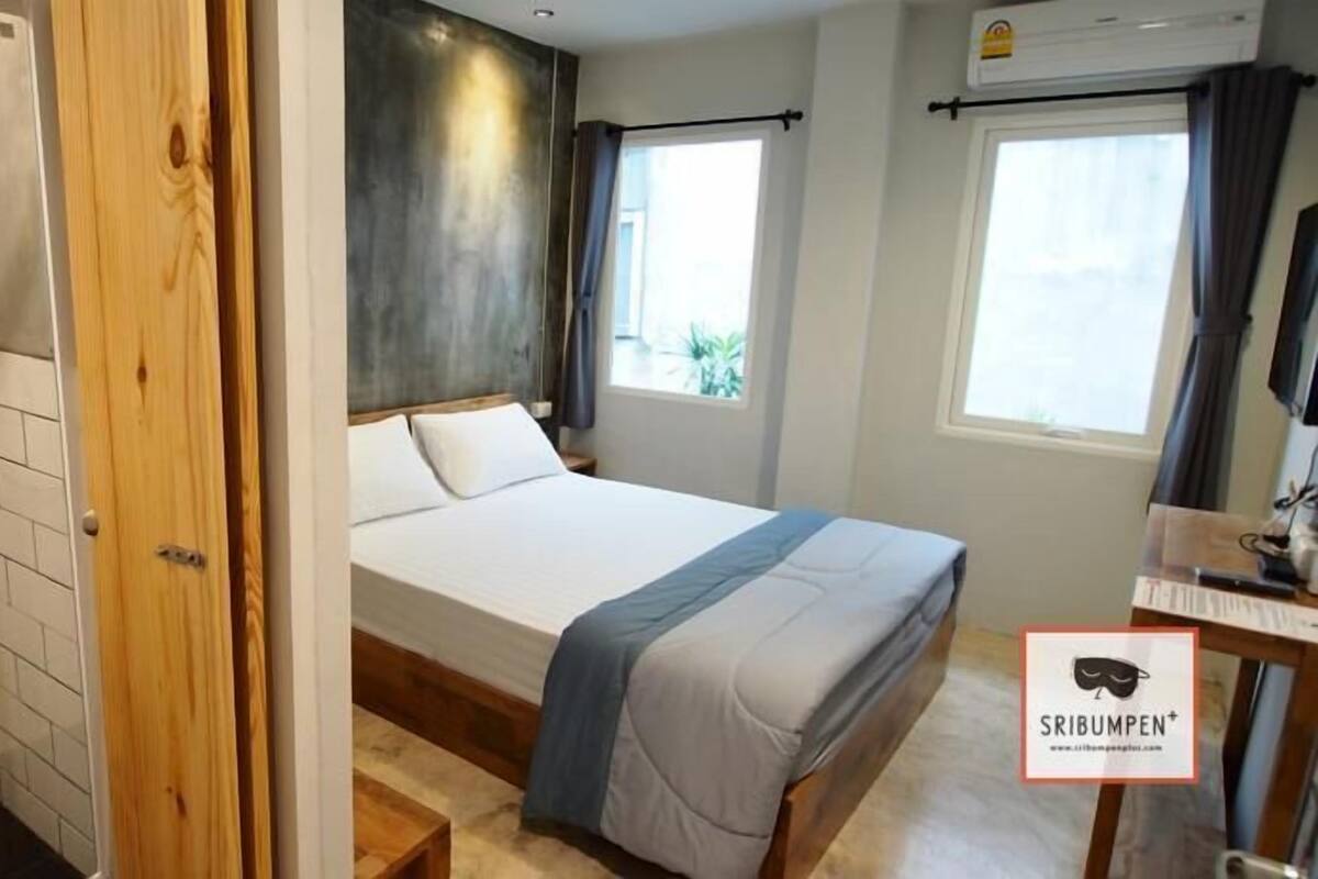A hotel room with a wooden door, a bed with a blue and white bedspread, a shower area with a white wall, and a window with curtains.