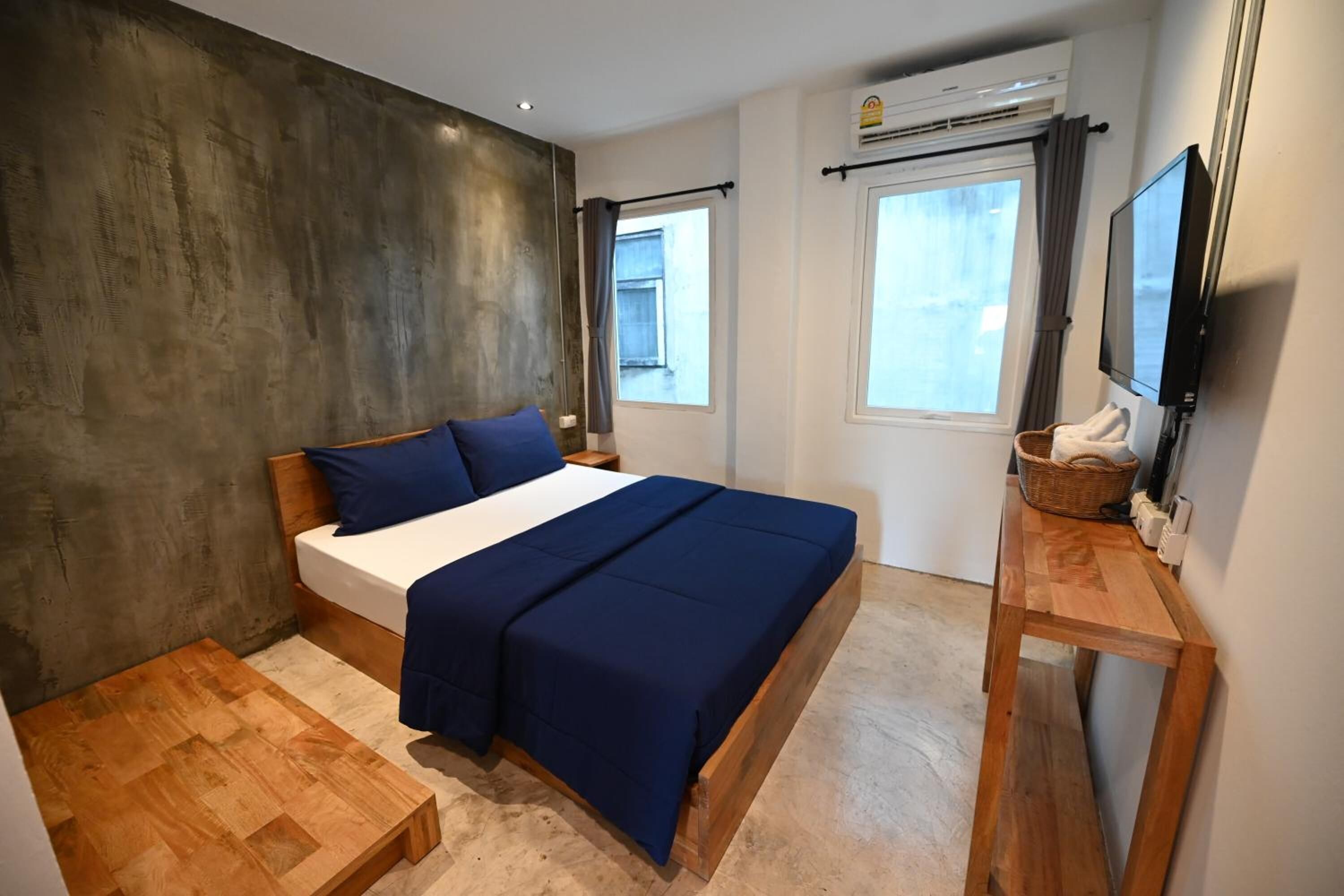 A bedroom with a bed, wooden floor, a small wooden table, a TV, and an air conditioning unit.