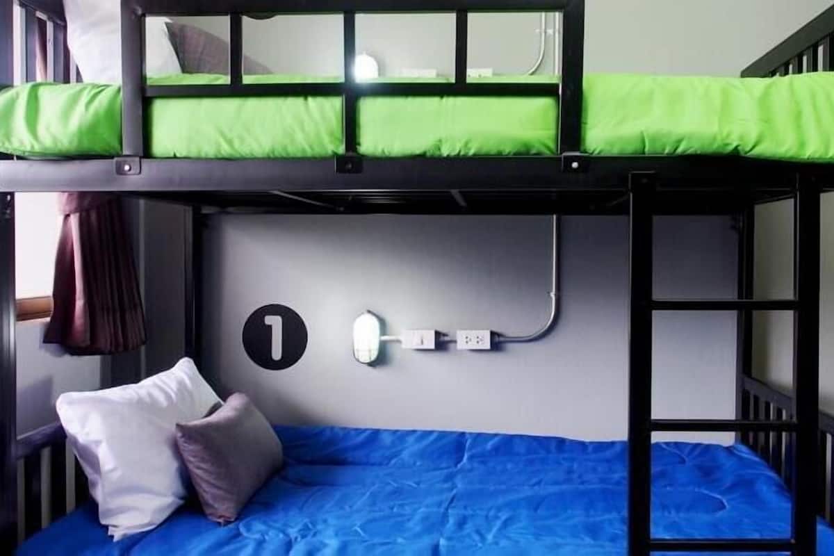 A bunk bed with a green mattress on the top bunk and a blue mattress on the bottom. There is a wall socket with a plug on the wall below the bed.