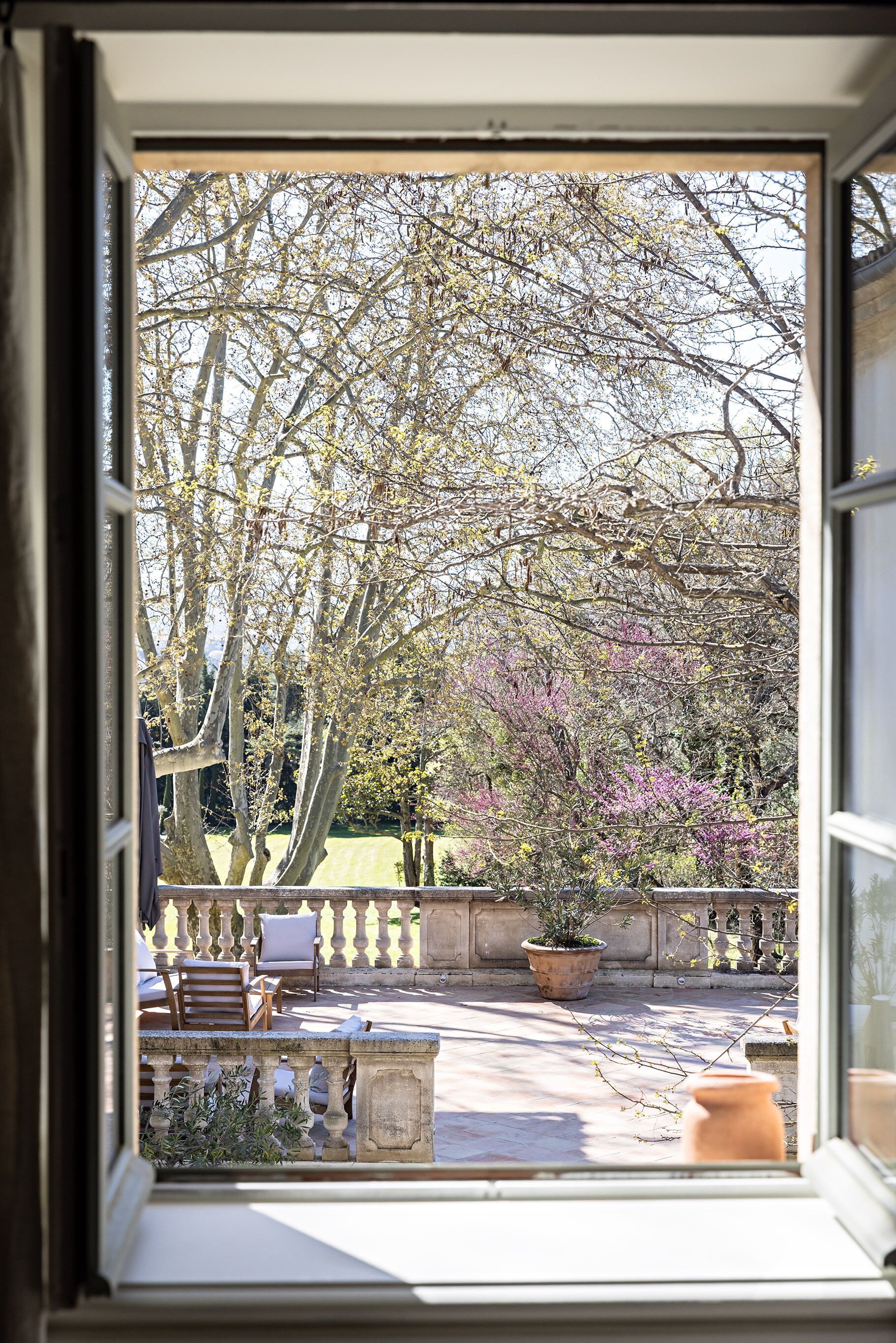 A view through a window of a courtyard with a bench, potted plants, and a stone wall.