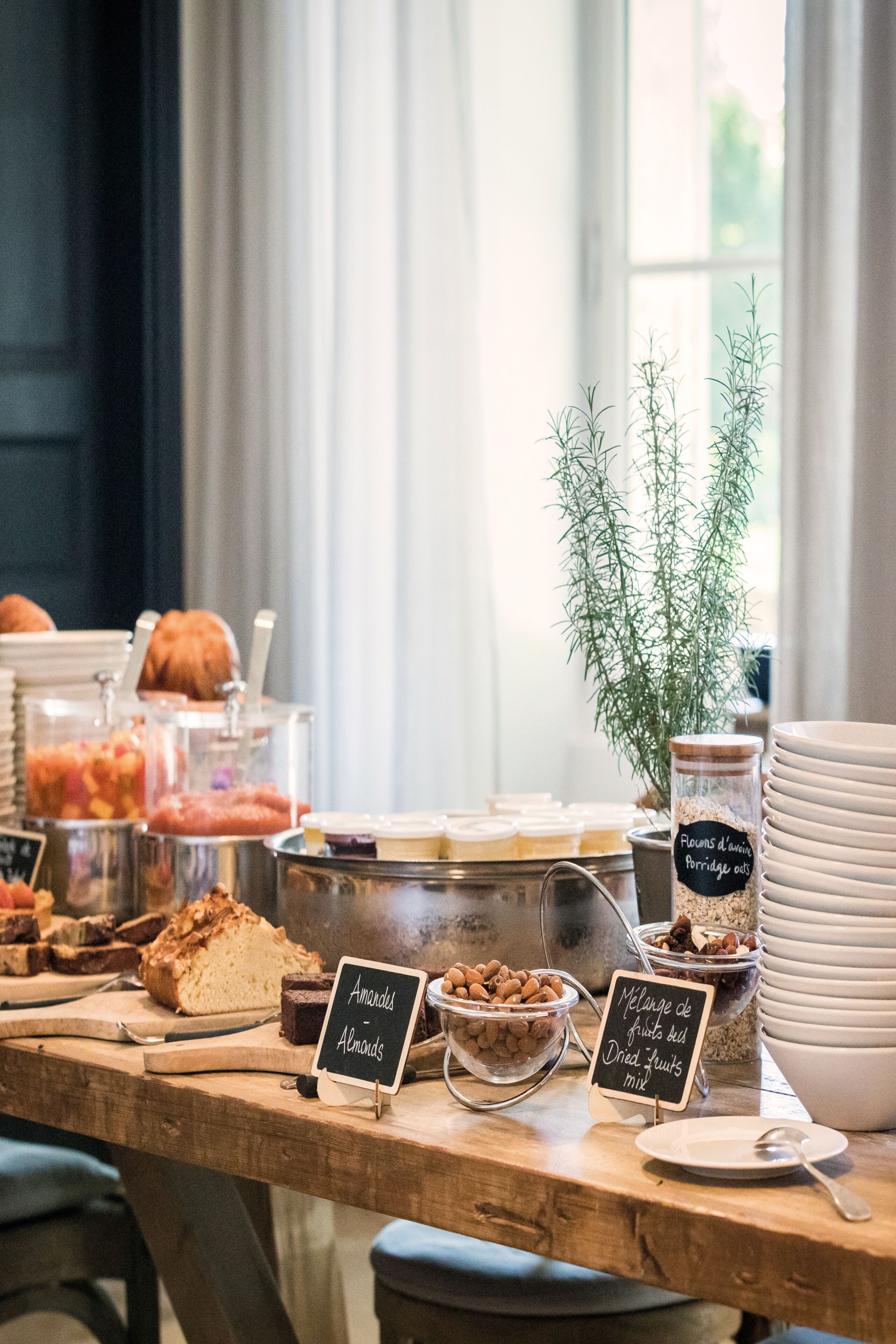 A breakfast buffet with various food items and labels in a bright room with a window.