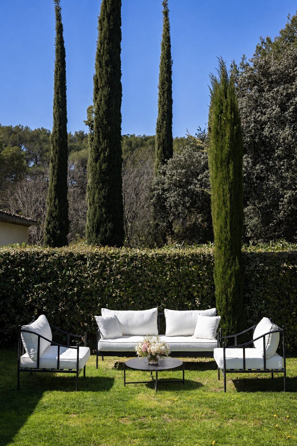 A garden with a white sofa set, a round coffee table, and tall cypress trees.