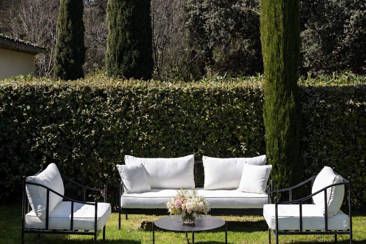 A garden with a white sofa set, a round coffee table, and tall cypress trees.