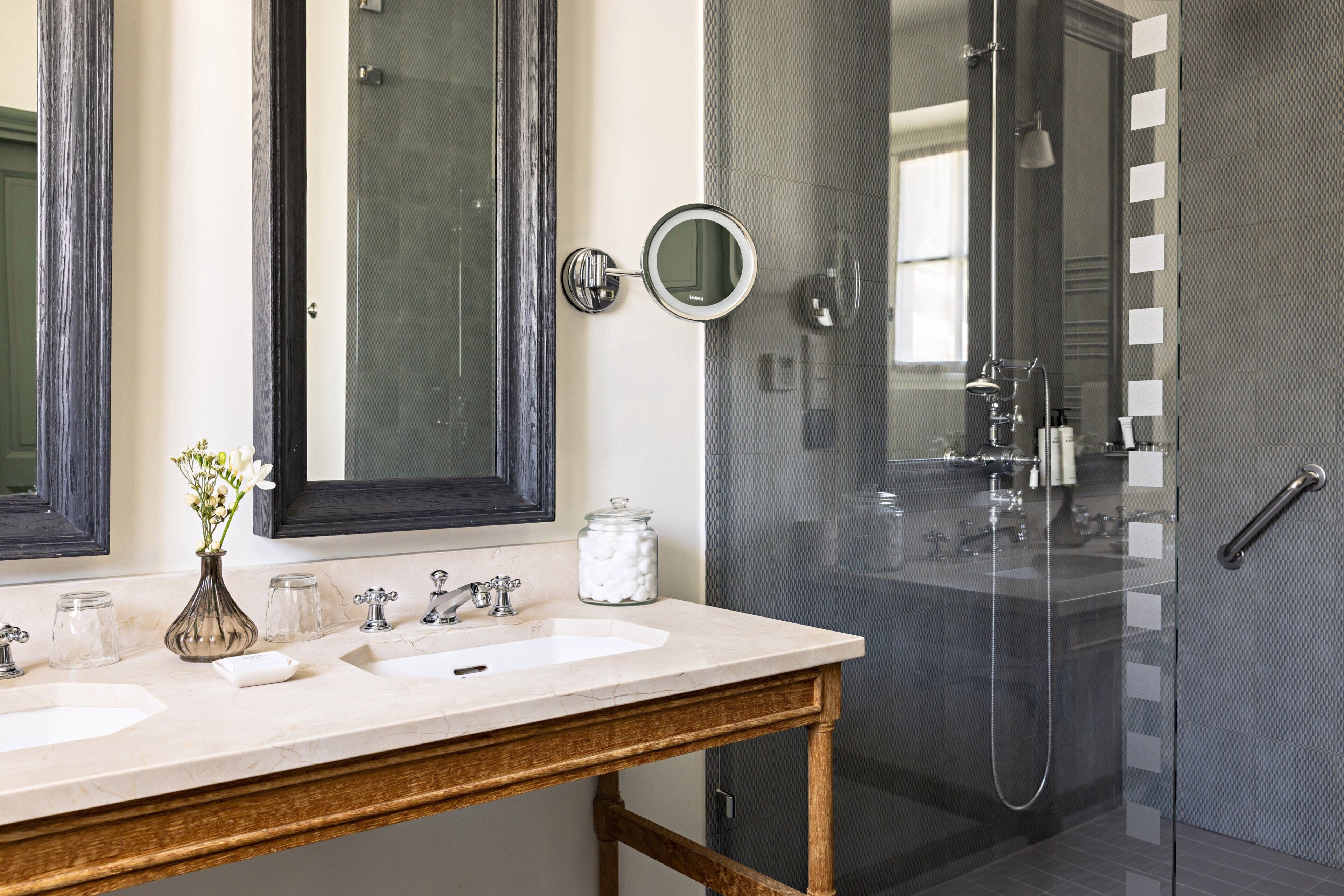 Une salle de bain avec deux lavabos, un miroir, un vase de fleurs et une cabine de douche en verre.