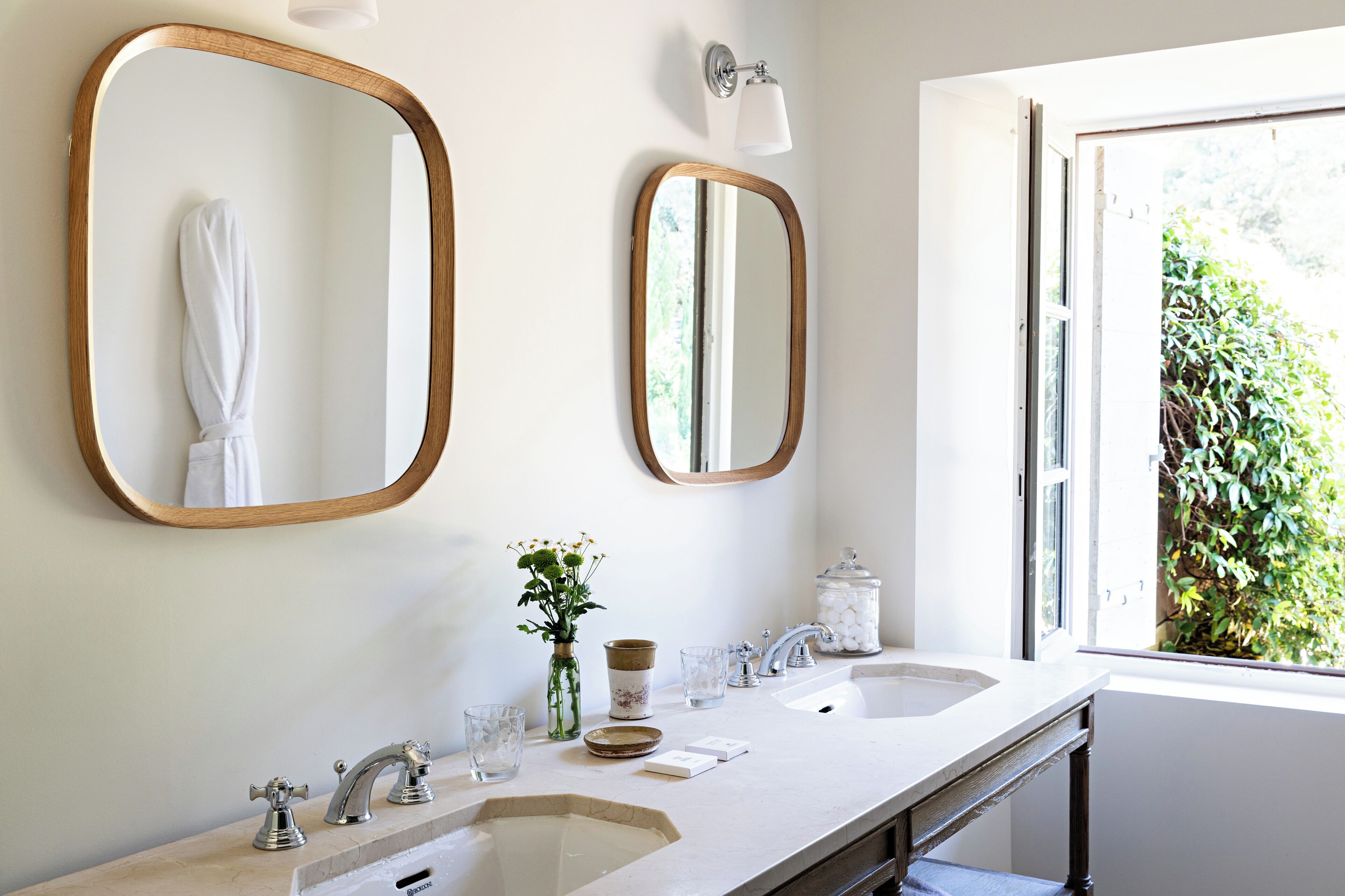 A bathroom with two sinks, a large mirror, a vase with flowers, and a window with a view of greenery.