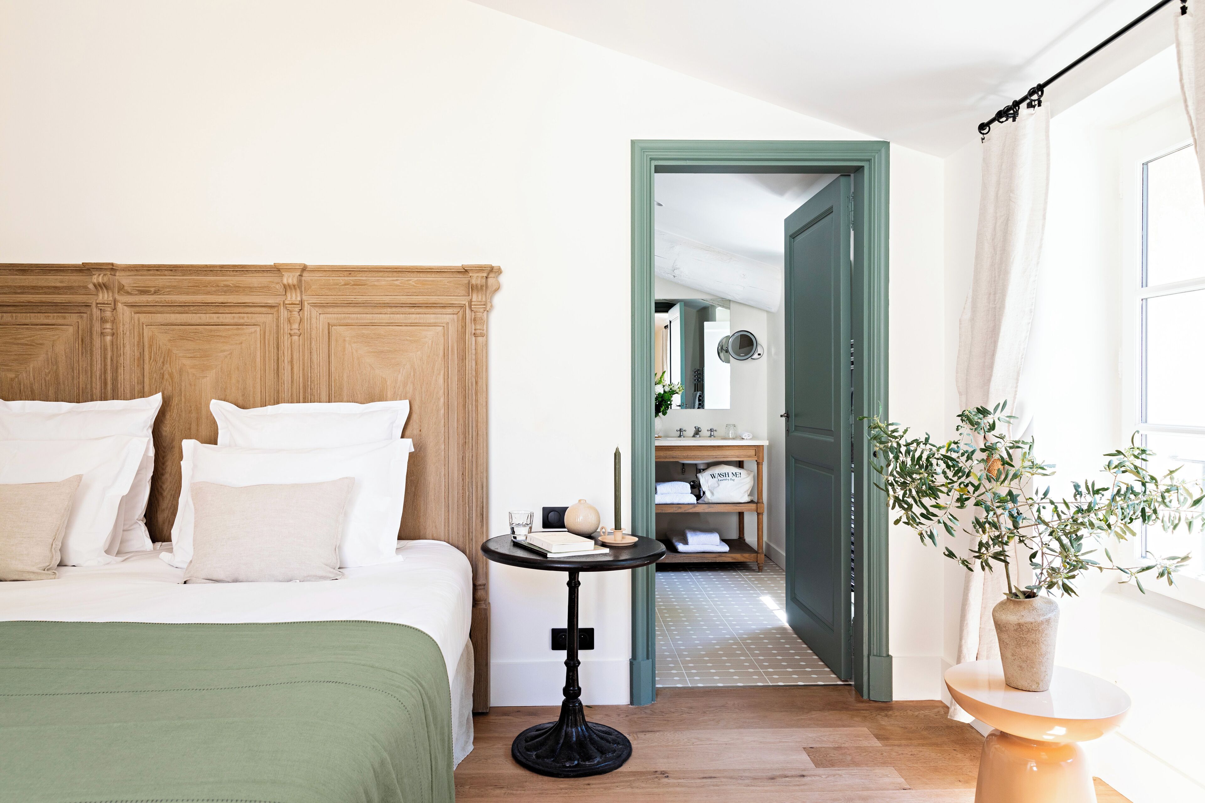 Un dormitorio con cama, una mesita de noche, una puerta verde y vistas a otra habitación con espejo y estanterías.