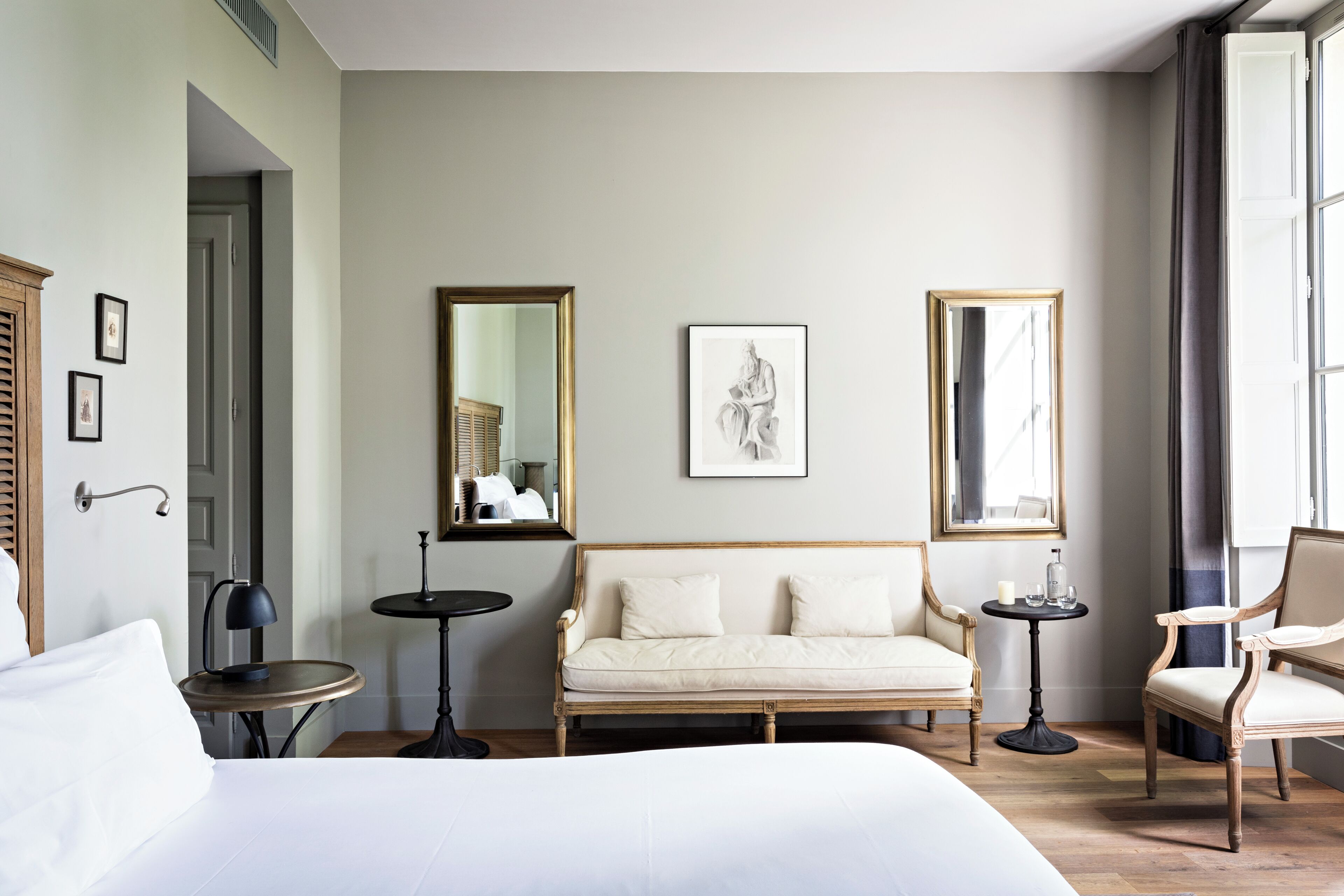Quarto de hotel com cama, sofá, duas cadeiras, uma mesinha e dois espelhos fixados na parede.