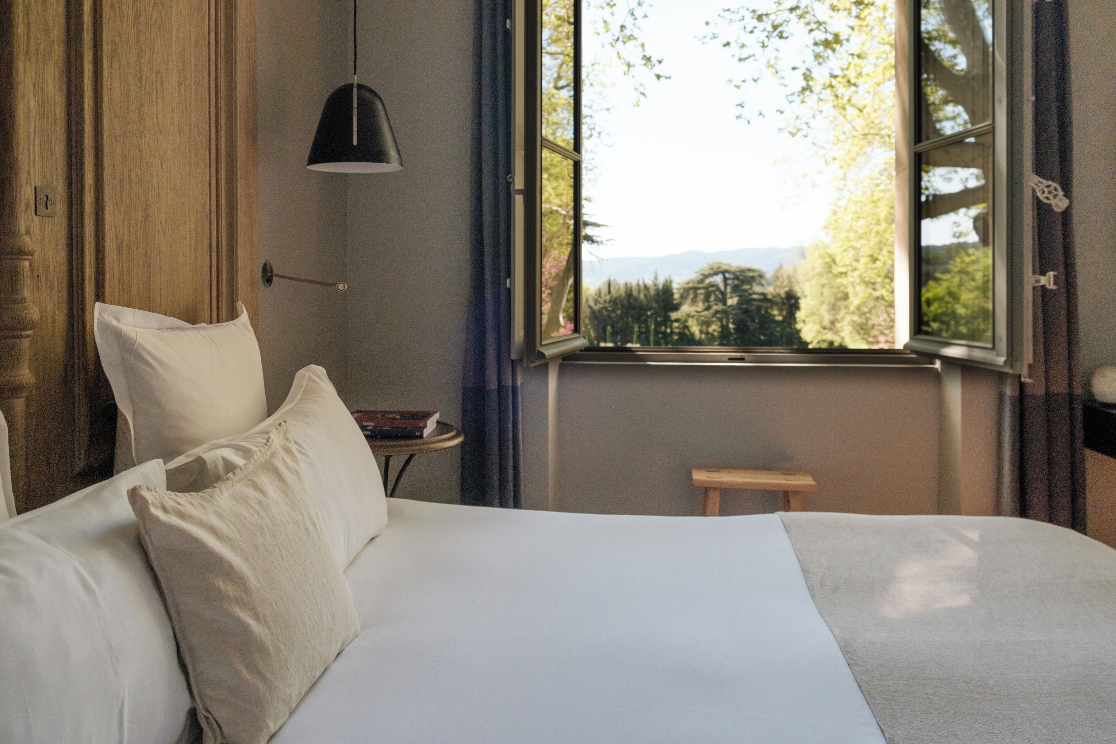 Une chambre à coucher avec un lit, une table de chevet sur laquelle se trouve un livre, une lampe et une fenêtre donnant sur des arbres et des montagnes.
